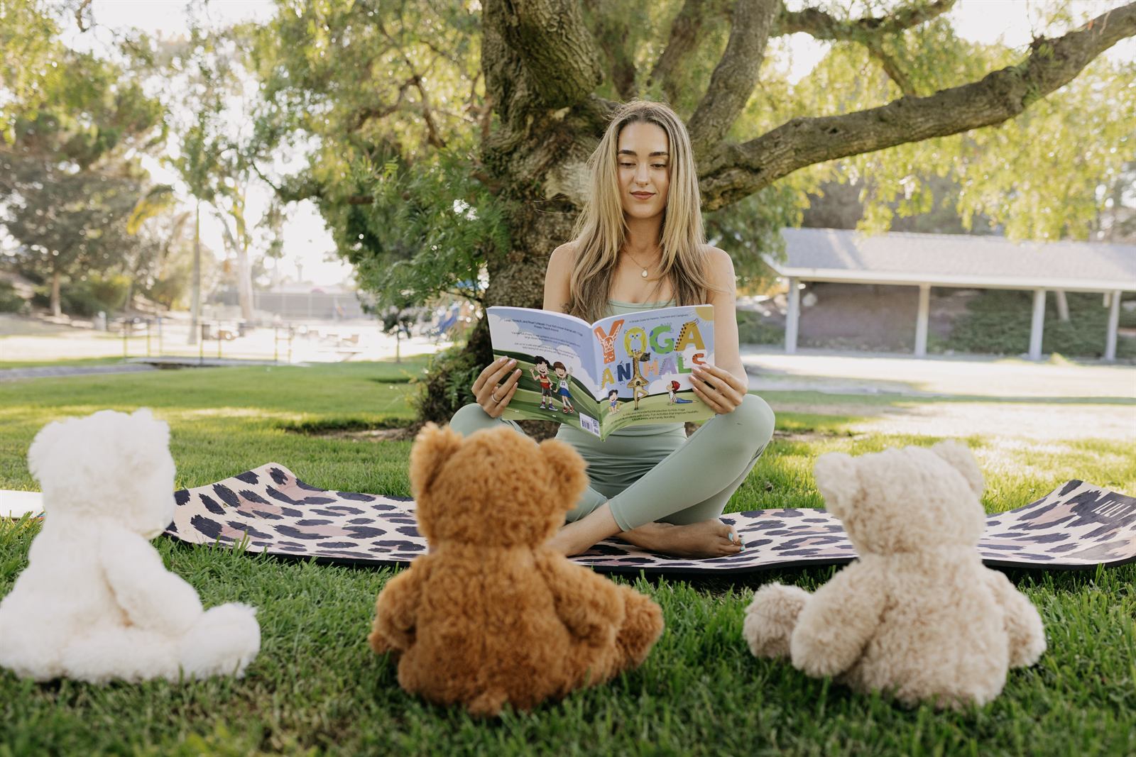 Teacher reading with three teddy bears on a mat under a tree